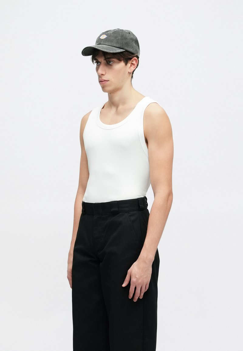 Hvid ribbede tanktop, sorte plisserede bukser og en mørk kasket med logo. Modellen står sidelæns mod en ensfarvet lys baggrund.