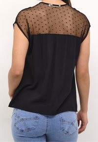 Haut noir avec un dos en tulle à pois, manches courtes et devant lisse. Associé à un jean bleu clair avec des poches arrière décoratives.