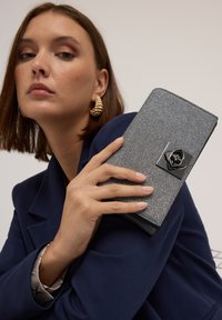 Glitzernde graue Clutch mit strukturierter Oberfläche, schwarzem Rosenakzent und elegantem Design, in einer Hand vor einem dunklen Blazer-Hintergrund gehalten.
