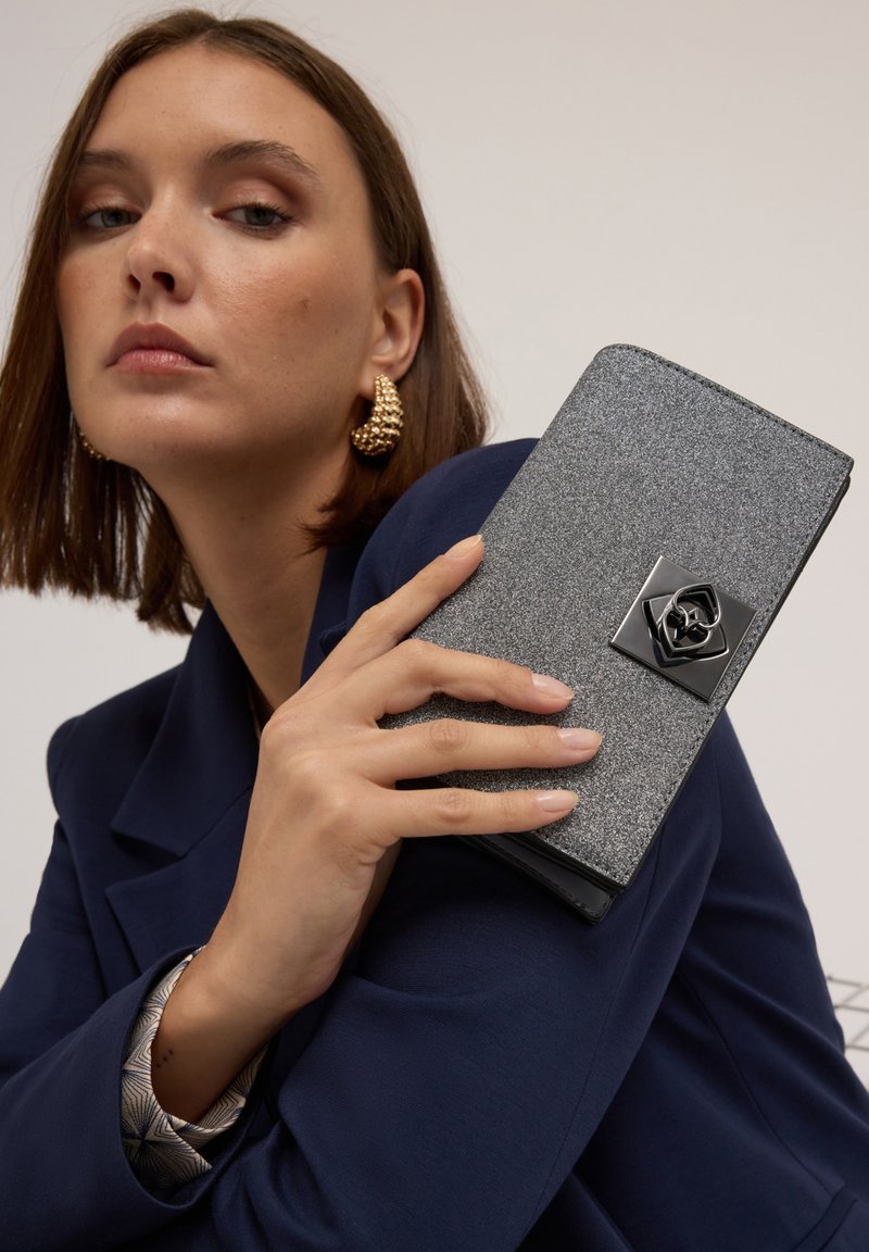 Glitzernde graue Clutch mit strukturierter Oberfläche, schwarzem Rosenakzent und elegantem Design, in einer Hand vor einem dunklen Blazer-Hintergrund gehalten.