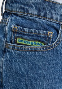 Modré džíny z denimu s zadnou kapsou, na ktorej je farebný vyšitý štítok v zelenej a modrej farbe s nápisom "DOSHOECO". Viditeľné stehy a detaily textúry.