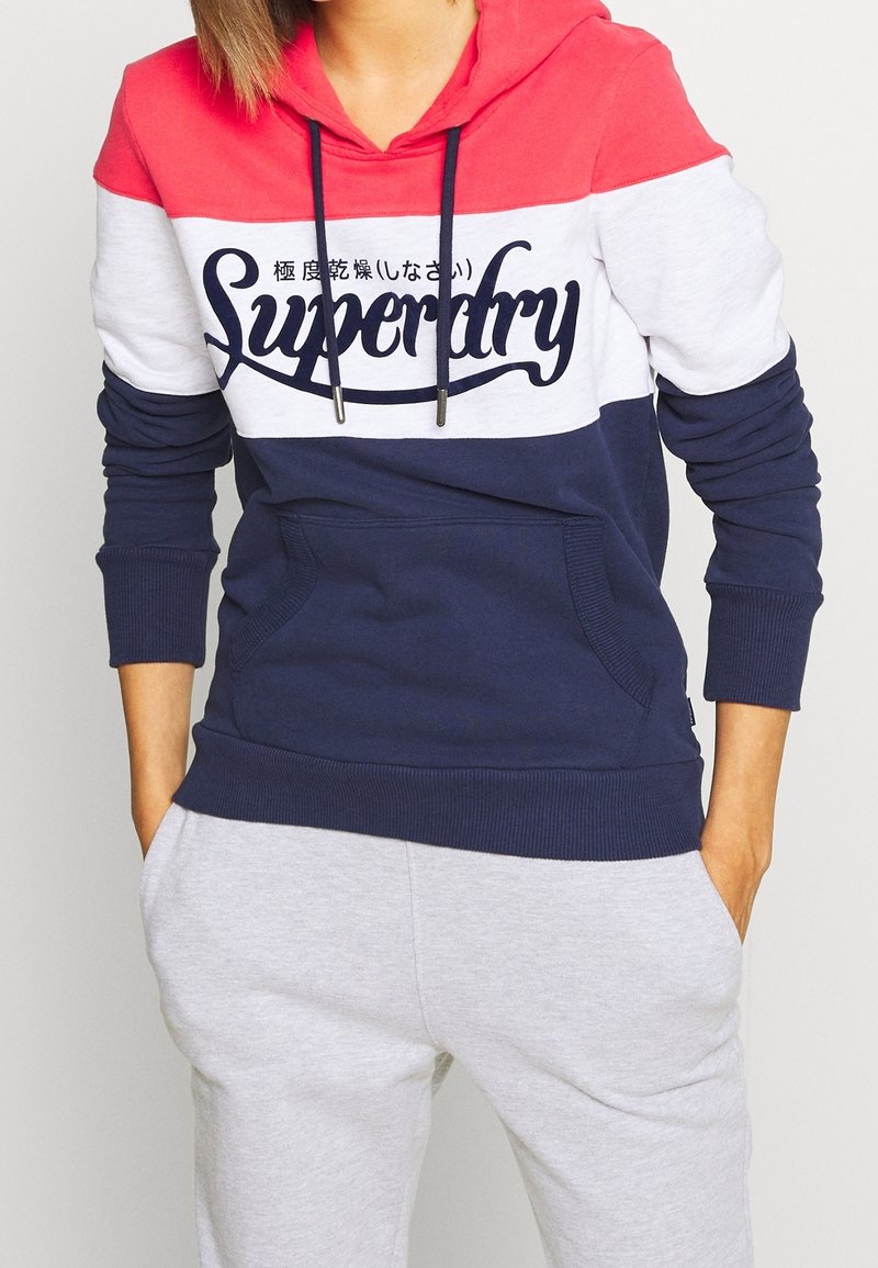 Personne portant un sweat à capuche color block avec des sections rouges, blanches et bleu marine, avec le texte "Superdry", assorti à un pantalon de survêtement gris clair.