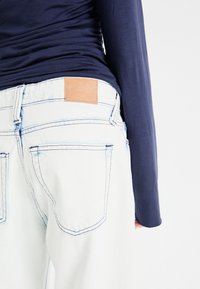 Jeans din denim albastru deschis cu cusături vizibile, o etichetă din piele maro pe talie și un top fitted din navy cu mânecă lungă parțial vizibil.