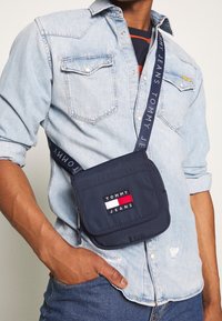 Borsa a tracolla blu navy con tracolla in tessuto che presenta il logo "TOMMY JEANS". La borsa ha una chiusura a pattina e una forma rettangolare.