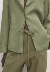 Personne portant une chemise boutonnée vert olive sur un pantalon à cordon assorti, une main dans la poche, cadrée au niveau du torse et des cuisses.