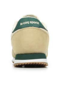 Tenisky s béžovým nubukovým svrchním materiálem, zelenými akcenty na patě, bílou mezipodešví a gumovou podešví; obsahují branding 'le coq sportif'.