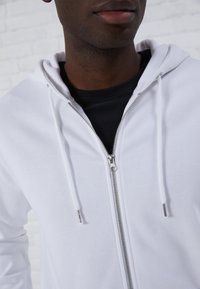 White zip-up hoodie with a smooth texture, silver zipper, and drawstrings, worn over a black shirt, against a light brick background.