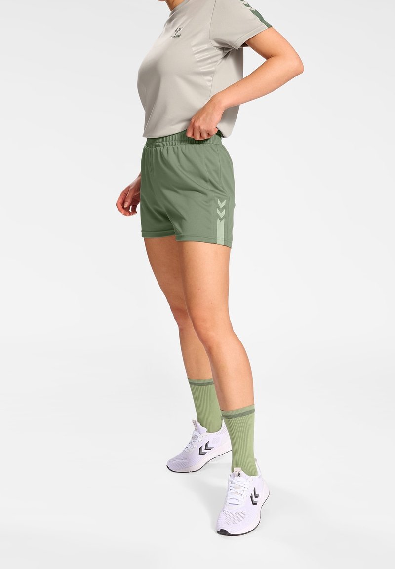 Pantalones cortos deportivos verdes con cintura elástica, combinados con una camiseta de manga corta gris claro, calcetines verdes y zapatillas blancas con detalles en negro.
