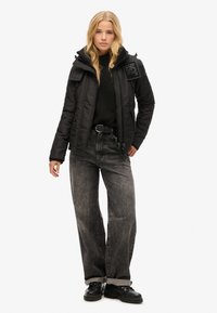 Veste rembourrée noire avec fermeture éclair à l'avant et capuche, associée à un jean gris taille haute et des bottes noires à lacets. Présence d'un logo sur la veste.