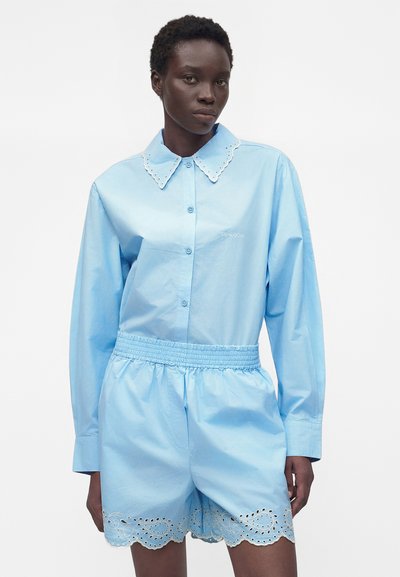 Γυναίκα που φοράει ανοικτό μπλε πουκάμισο με κουμπιά και γιακά με κυματιστά άκρα και αντίστοιχα ελαστικά σορτς με δαντέλα με οπές.