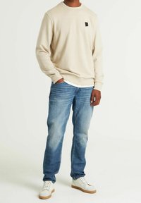 Felpa beige a girocollo, vestibilità rilassata, con un piccolo patch logo nero sul petto. Abbinata a jeans azzurri chiari e sneaker bianche.