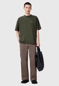 Camiseta oversized verde oliva, pantalones cargo beige, zapatos negros tipo slip-on y un bolso mensajero oscuro; ajuste relajado con detalles mínimos.