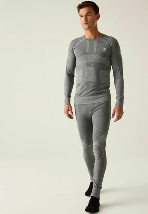 Homme portant un haut thermique gris ajusté à manches longues et des leggings, debout sur un fond clair uni avec des chaussettes noires.