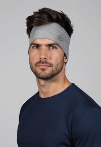 Grijze sportieve hoofdband gemaakt van zachte, rekbare stof, gedragen over een donkerblauw shirt. Voorzien van een logo aan de zijkant en een gladde textuur.