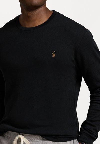 Čierne tričko s dlhými rukávmi vyrobené z mäkkej látky, s malým vyšitým logom polo na hrudi, v kombinácii so svetlo šedými nohavicami na šnúrku.