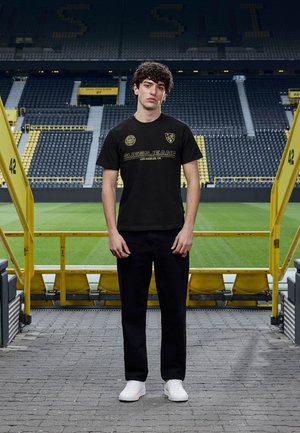 Junger Mann mit lockigem Haar steht nach vorne gerichtet auf den Stadionstufen, trägt ein schwarzes Guess Jeans Hemd und Hose mit weißen Sneakers.