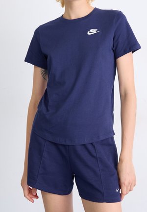 Persoon draagt een marineblauw Nike T-shirt en bijpassende korte broek met witte Nike-logo's op de borst en korte broek tegen een effen achtergrond.