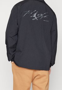 Chaqueta negra con tejido texturizado, capucha, puños ceñidos y un logo reflectante de 'Flight' en la parte trasera. Diseño informal.