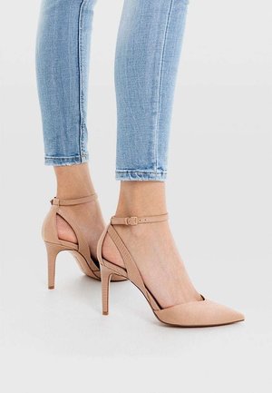 Pieds portant des talons aiguilles couleur nude à bouts pointus avec des brides à la cheville, associés à un jean skinny bleu clair sur un fond uni.