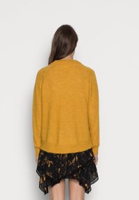 Frau mit langen braunen Haaren, die einen senfgelben Pullover und einen schwarzen, floral gemusterten, gestuften Rock trägt, mit dem Rücken zu einem schlichten weißen Hintergrund.