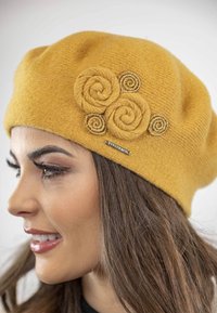 Żółty wełniany beret z trzema akcentami w kształcie spiralnych kwiatów w beżu i złotych rzemykach, z małą metalową metką z logo. Gładka tekstura.