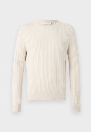 Homme aux cheveux bouclés et foncés portant un pull en maille torsadée crème, un jean bleu large et des baskets blanches, debout avec une main dans la poche.