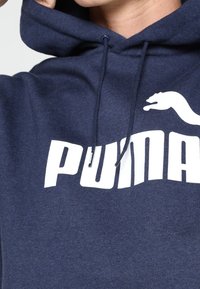 Felpa con cappuccio blu navy realizzata in tessuto morbido, con un grande logo "PUMA" bianco e un piccolo dettaglio logo bianco laterale. Cappuccio regolabile con cordino.