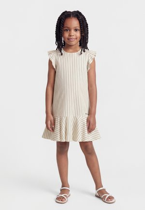 Mladá dívka s copánky, oblečená v krémových bezrukávých šatech s tenkými vertikálními pruhy a bílých sandálech, stojí před jednobarevným pozadím.