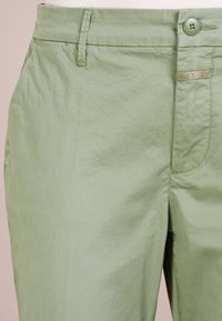 Pantaloni di cotone verde chiaro con una consistenza liscia, dotati di chiusura a bottone, tasche laterali e dettagli cuciti sulla vita.