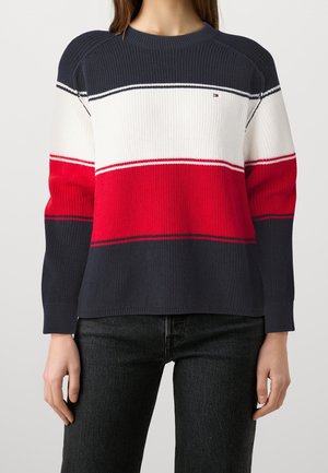 Femme portant un pull en maille rayé bleu marine, blanc et rouge à manches longues et un pantalon noir.