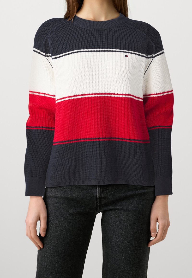 Femme portant un pull en maille rayé bleu marine, blanc et rouge à manches longues et un pantalon noir.