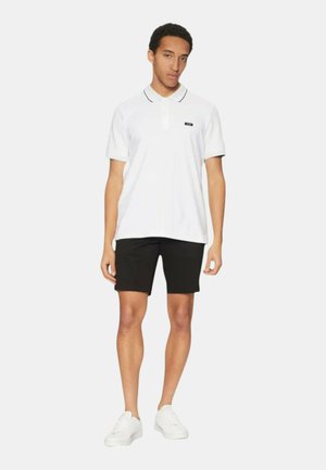 Polo shirt bianca con logo nero, caratterizzata da un colletto con bordi neri, abbinata a pantaloni corti neri e sneakers bianche. Design semplice e pulito.