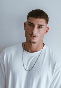 Tricou alb din bumbac, cu mâneci scurte, design simplu și guler rotund. O brățară din lanț negru se află pe piept, fundal simplu.