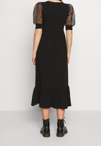Musta midi-mekko, jossa on puffimaiset läpinäkyvät hihat, rypistetty vyötärö ja levenevä helma. Yhdistetty mustiin nilkkabootseihin. Näkyy takaa.