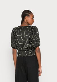 Blusa preta com padrão, featuring mangas bufantes, volantes texturizados e um design de linha ondulada branca com acabamentos metálicos.