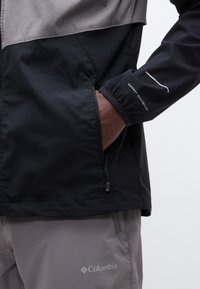 Χέρι ατόμου στην τσέπη ενός μαύρου και γκρι μπουφάν με φερμουάρ, φορά γκρι παντελόνι Columbia με εμφανή λογότυπα στο μανίκι του μπουφάν και το παντελόνι.