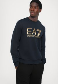 Navy blue sweatshirt made of cotton blend, featuring a gold logo print. Crew neck, ribbed cuffs, and hem with a smooth, soft texture.