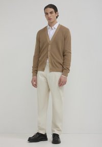 Cardigan lavorato a maglia beige con cinque bottoni, abbinato a pantaloni crema e scarpe nere, in piedi davanti a uno sfondo neutro.
