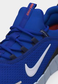 Κοντινό πλάνο μπλε αθλητικού παπουτσιού Nike Free που δείχνει τον πλεκτό επάνω μέρος, το λευκό λογότυπο Swoosh, τα μπλε κορδόνια και την ταινία έλξης με το κείμενο "NIKE FREE" σε πορτοκαλί.