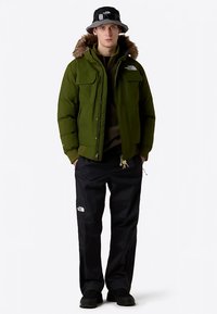 Zelená péřová bunda s kapucí lemovanou kožešinou, dvěma kapsami na hrudi, černé kalhoty s logem a černý klobouk typu bucket se silnou podrážkou.