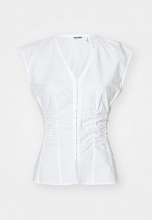 Blouse en coton blanc avec des manches courtes type cap, design ajusté, décolleté en V, fermeture à boutons sur le devant et détails froncés sur les côtés.