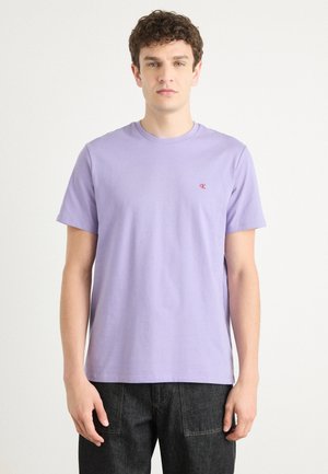Camiseta de algodón de manga corta en color lavanda, con cuello redondo y pequeño logo rojo en el lado izquierdo del pecho. Combinada con jeans oscuros.