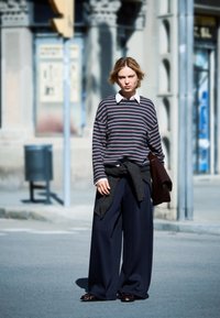 Camicia a righe con maniche lunghe e colletto bianco, abbinata a pantaloni larghi scuri. Un maglione grigio è annodato in vita. Sono indossate scarpe marroni.