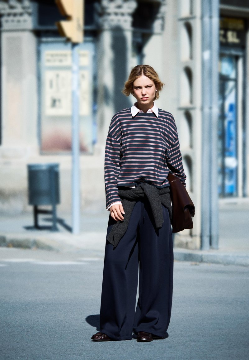 Prugasta košulja dugih rukava s bijelim ovratnikom, uparena s labavim tamnim hlačama. Siva majica je zavezana oko struka. Nosi se smeđa obuća.