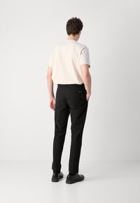 Pantaloni neri slim-fit con una texture liscia, indossati con una camicia beige a maniche corte. Le scarpe sono nere, con una suola bassa e un design minimale.