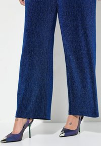 Pantaloni a gamba larga di colore blu con texture, abbinati a decolletè con tacco a gradiente viola, dotati di punte metalliche e tacchi a spillo verdi.