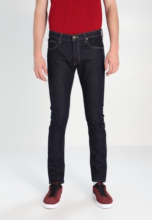 Jeans de mezclilla azul oscuro con un ajuste slim, que presentan costuras amarillas, cinco bolsillos y una cremallera. Combinados con zapatillas rojas.