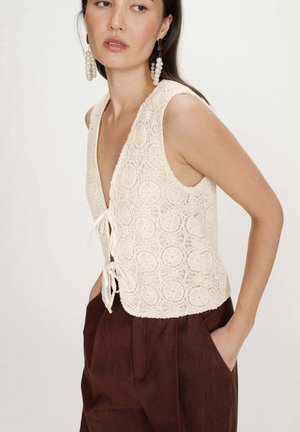 Femme portant un haut en crochet crème sans manches avec des liens et un pantalon marron, accessoirisée de grandes boucles d'oreilles en perles, posant de profil.