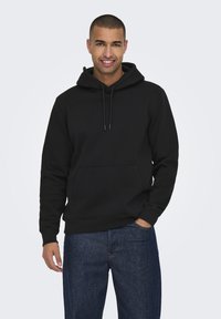 Felpa nera con cappuccio realizzata in un morbido e resistente tessuto elasticizzato. Presenta una tasca frontale, cordini e polsini a coste. Indossata con jeans blu.