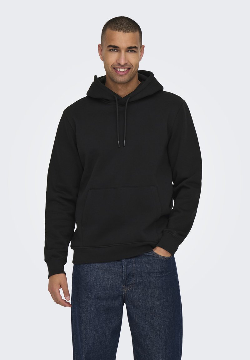 Felpa nera con cappuccio realizzata in un morbido e resistente tessuto elasticizzato. Presenta una tasca frontale, cordini e polsini a coste. Indossata con jeans blu.